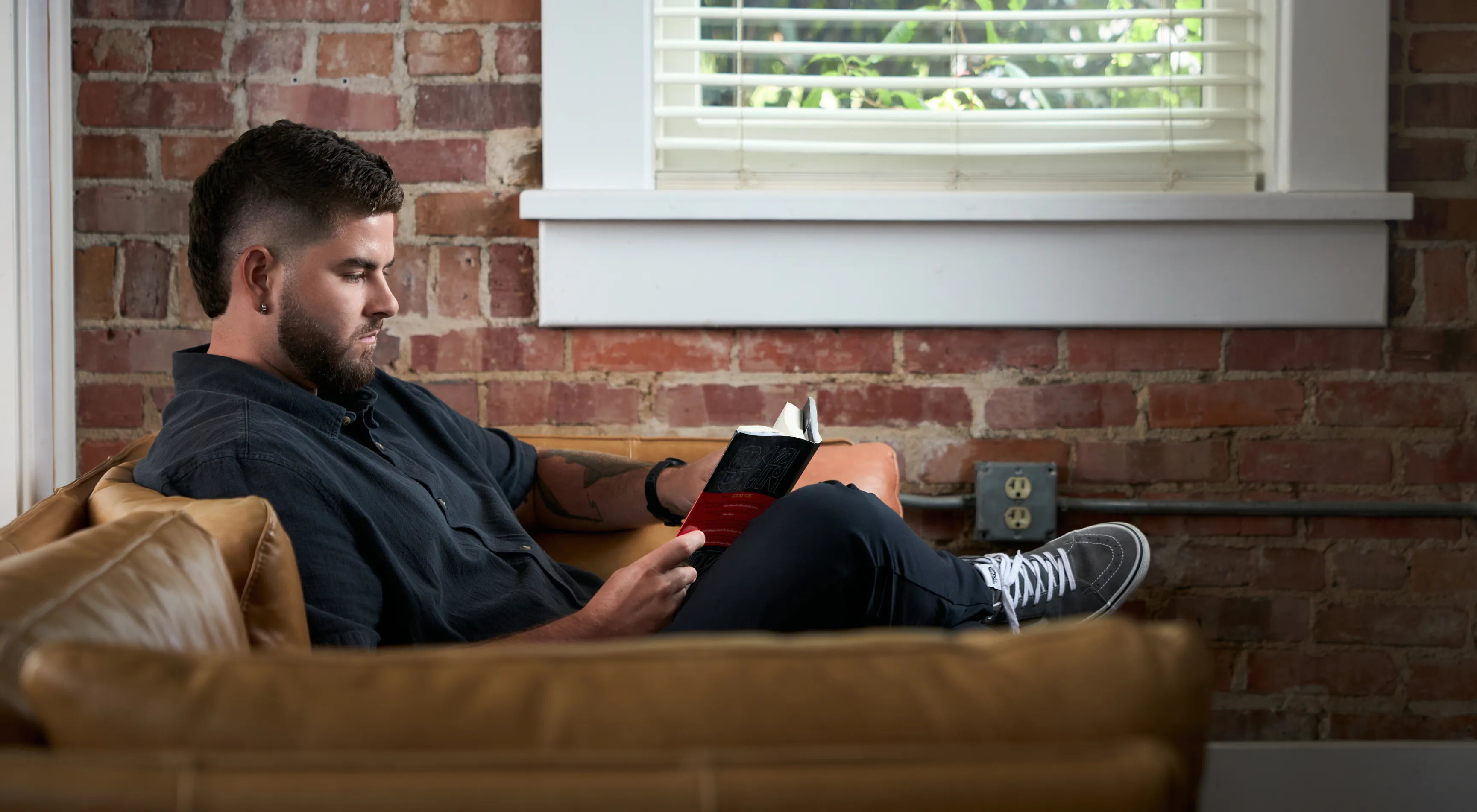 Unbound Collective team member reading a book on a leather sofa in the studio against an exposed brick wall, brand photography by Unbound Collective