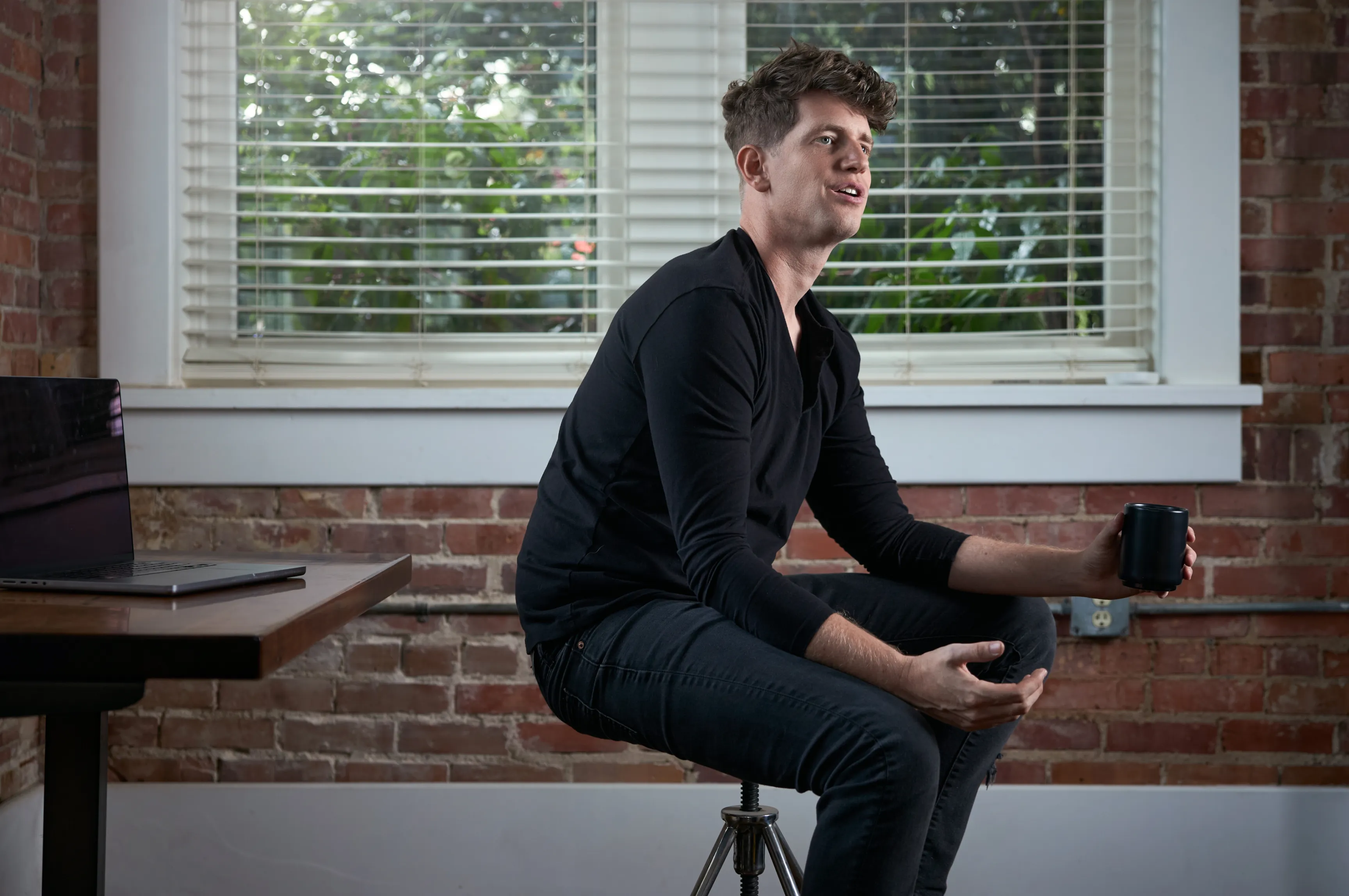 Unbound Collective Design Strategist Hunter Oden seated on a stool holding a coffee mug in the studio against an exposed brick wall, brand photography by Unbound Collective