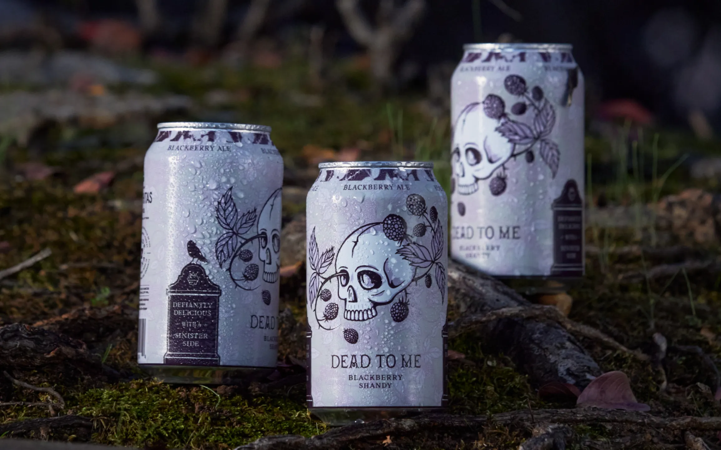 The Ouachitas Brewing Dead to Me Blackberry Shandy cans with skull and blackberry illustration resting on moss and roots, packaging design by Unbound Collective