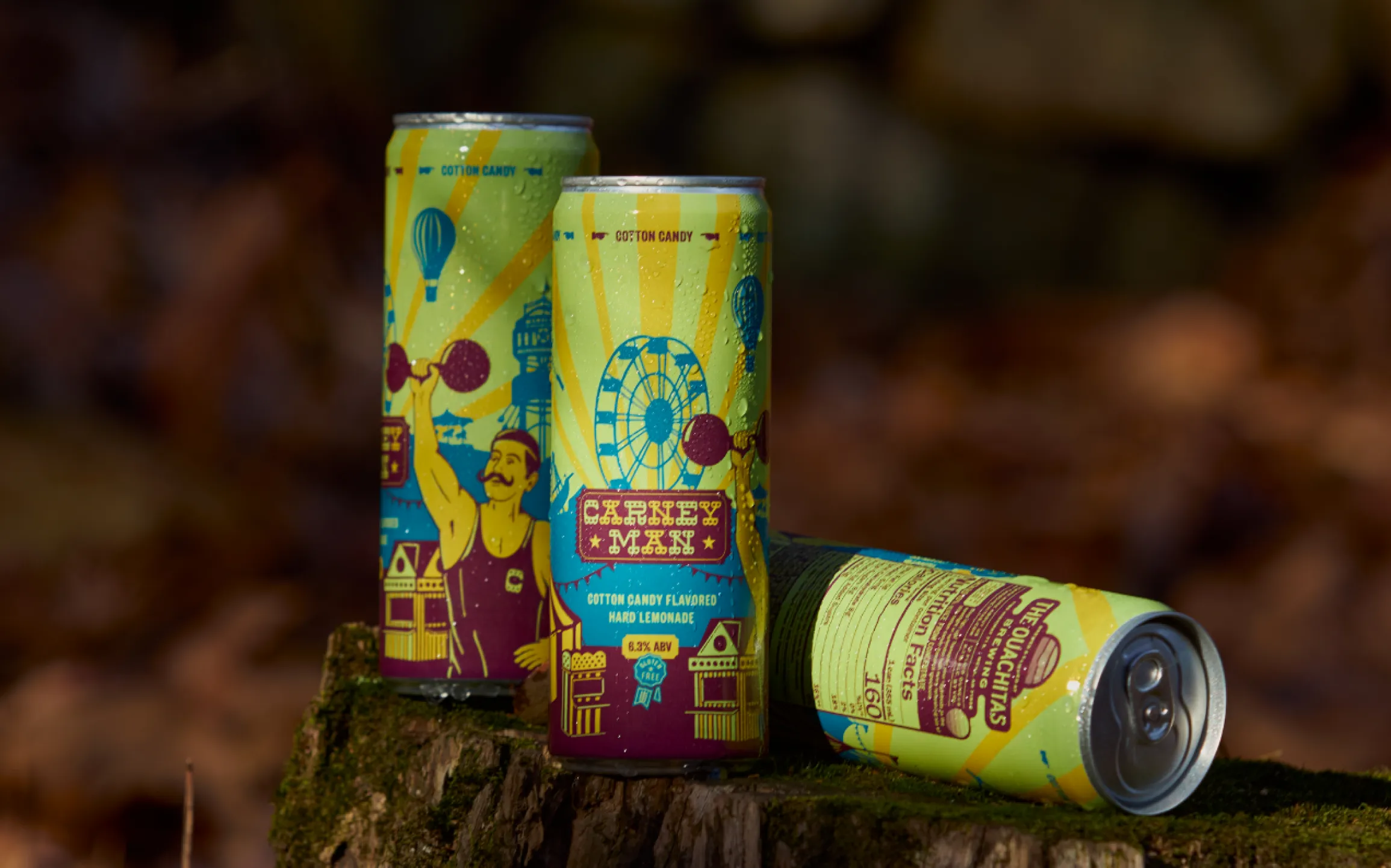 The Ouachitas Brewing Carney Man Cotton Candy Hard Lemonade tall cans with carnival strongman and ferris wheel illustration on a mossy stump, packaging design by Unbound Collective