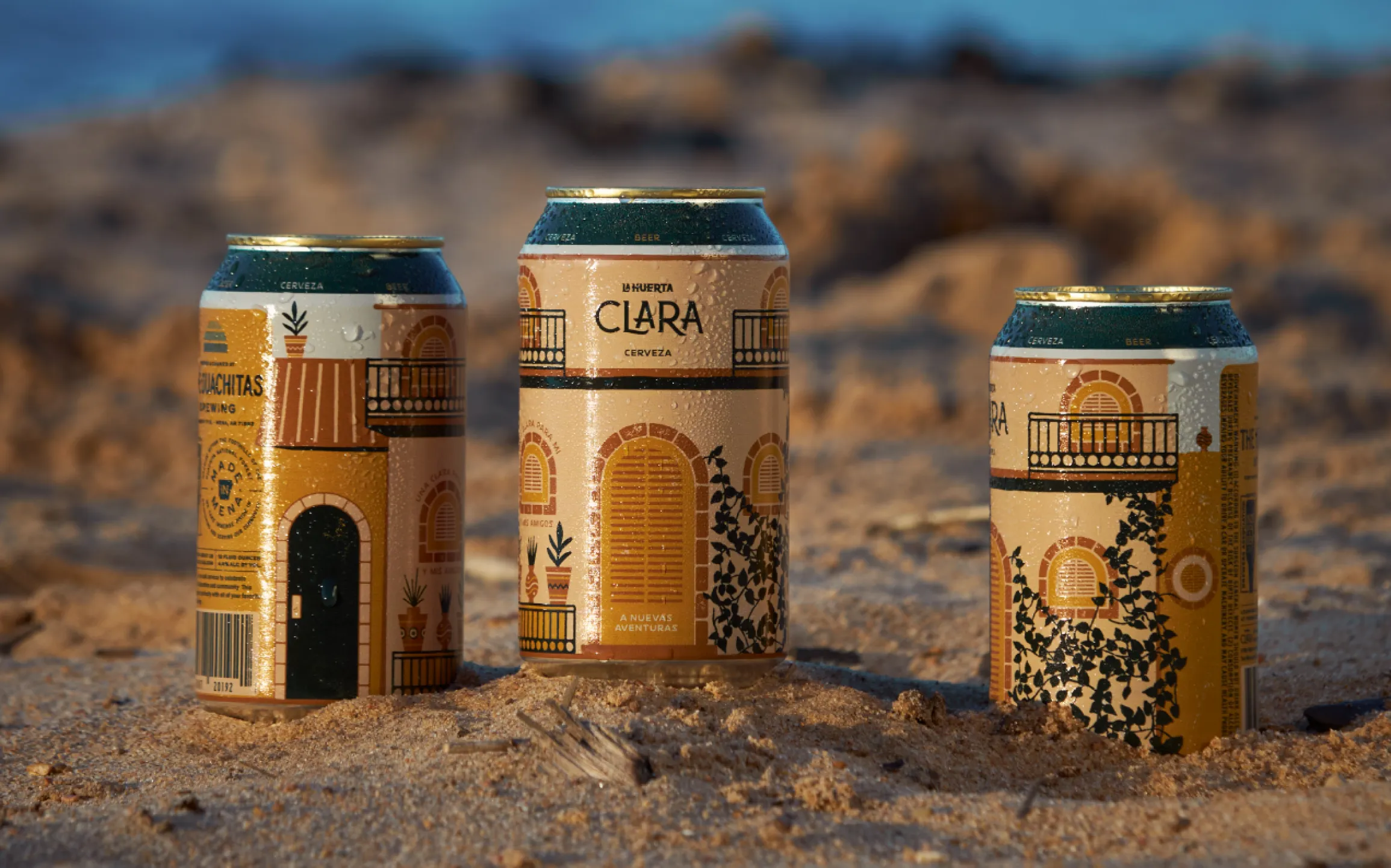 The Ouachitas Brewing La Huerta Clara Cerveza cans with illustrated hacienda facade design standing in beach sand, packaging design by Unbound Collective