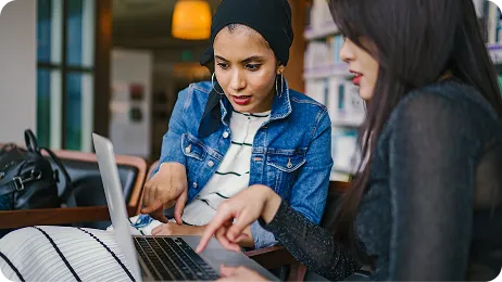 Two young women collaborating on a laptop in a library or café, pointing at the screen while discussing work.
