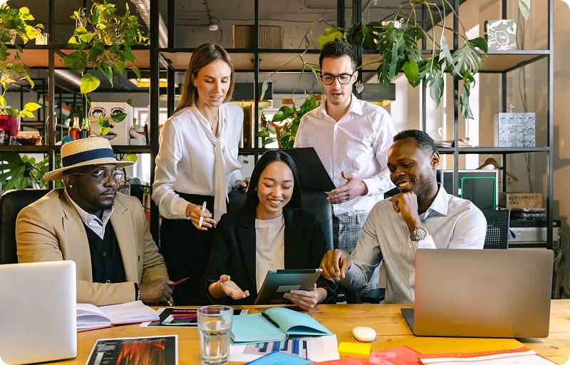 A diverse group of five professionals collaborating around a table in a modern office, reviewing documents and a tablet while discussing ideas.
