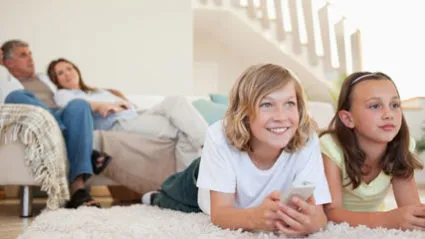 A family scene with two children and two adults. The image shows two children lying on a carpet, watching TV or playing with a remote, while two adults are seated on a couch in the background.