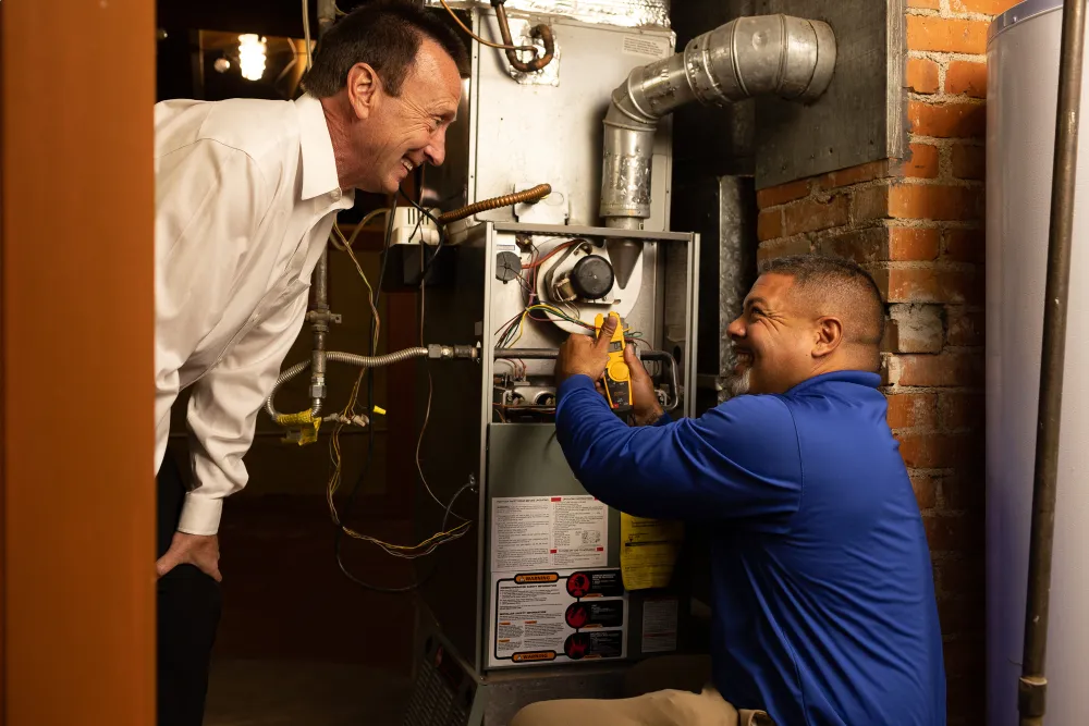 A technician in a blue Wagner shirt is working on a furnace in a basement, using tools to check the connections. An older man in a white shirt is leaning in, watching the technician with interest and smiling