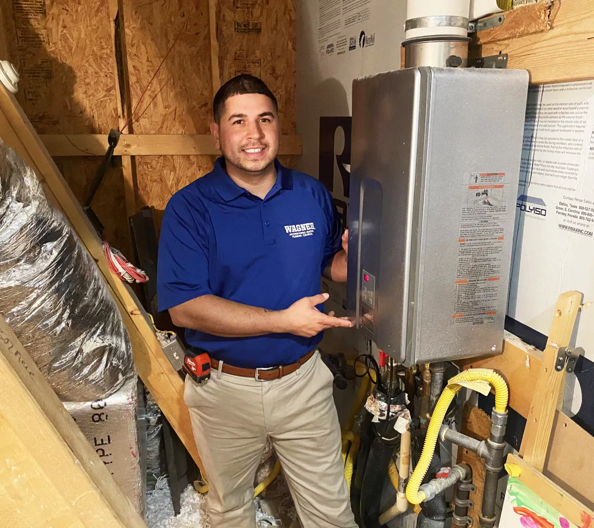 A technician in a blue Wagner shirt is smiling and pointing at a tankless water heater installed in a utility room. The room has exposed wooden beams, ductwork, and various instructional labels on the walls.