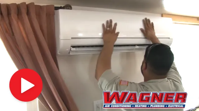 A technician is standing on a ladder and installing a wall-mounted air conditioning unit. The technician, wearing a light-colored shirt with an emblem, is reaching up to secure the unit. In the background, there are beige curtains partially drawn, and a wall with electrical outlets is visible. At the bottom left, there is a red play button overlaid indicating that the image is a video. The Wagner company logo, which offers air conditioning, heating, plumbing, and electrical services, is prominently displayed at the bottom.