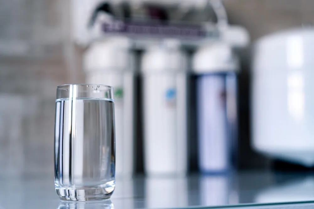 A clear glass filled with water sits on a reflective surface in the foreground. Behind the glass, there are several water purification or filtration devices with different colored sections, indicating a domestic water filtration system. The background is softly blurred, drawing attention to the glass of water in the front. The overall setting suggests a clean, modern kitchen environment.