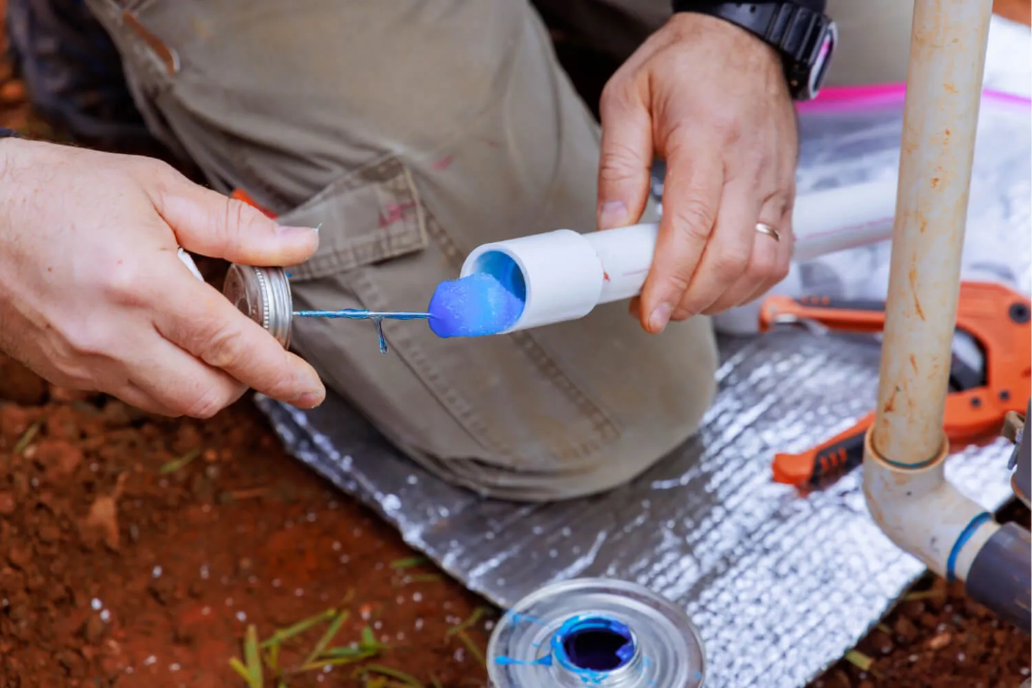 A person is applying a blue adhesive or sealant into a white PVC pipe using a small tool held in their left hand. The right hand is positioned to hold the PVC pipe steady. The background consists of reddish-brown soil and a silver reflective surface, with tools visible nearby, including a pipe cutter and clamps. The person is wearing light-colored pants with pockets.