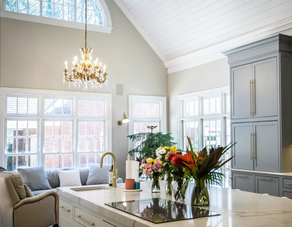 Spacious kitchen with high ceilings, chandelier, large windows, and an island with flowers in vases.