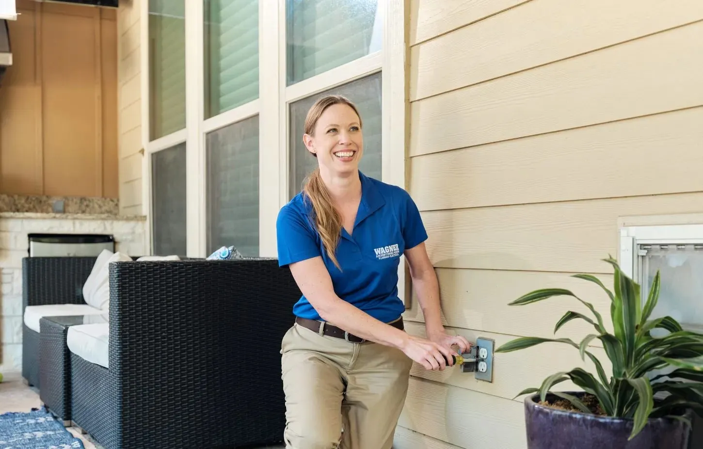 A person in a blue shirt and khaki pants is smiling and adjusting a pipe on the exterior wall of a house, next to a potted plant.