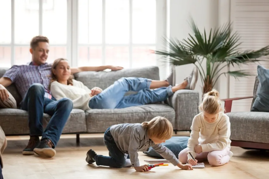 Two children sitting on the floor drawing with markers while two adults relax on a gray couch in the background of a bright living room.