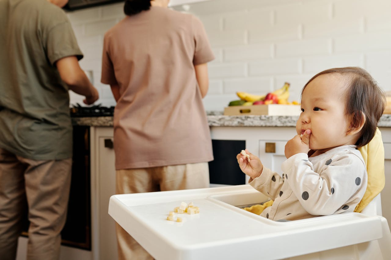 baby sitting in a high chair in the kitchen eating with parents cooking at the counter in the background.