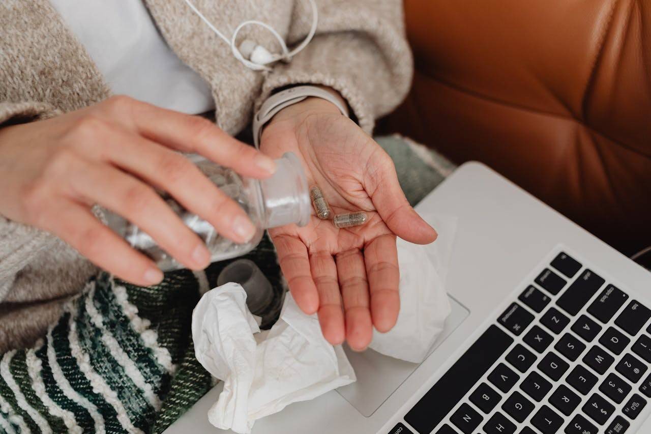 Person pouring capsules into their hand beside a laptop, with tissues and earphones nearby.