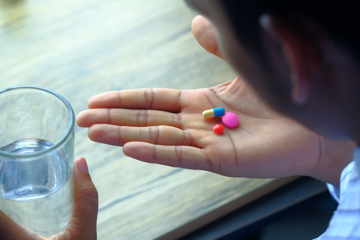 A person holds three colorful pills in their palm next to a glass of water on a wooden table