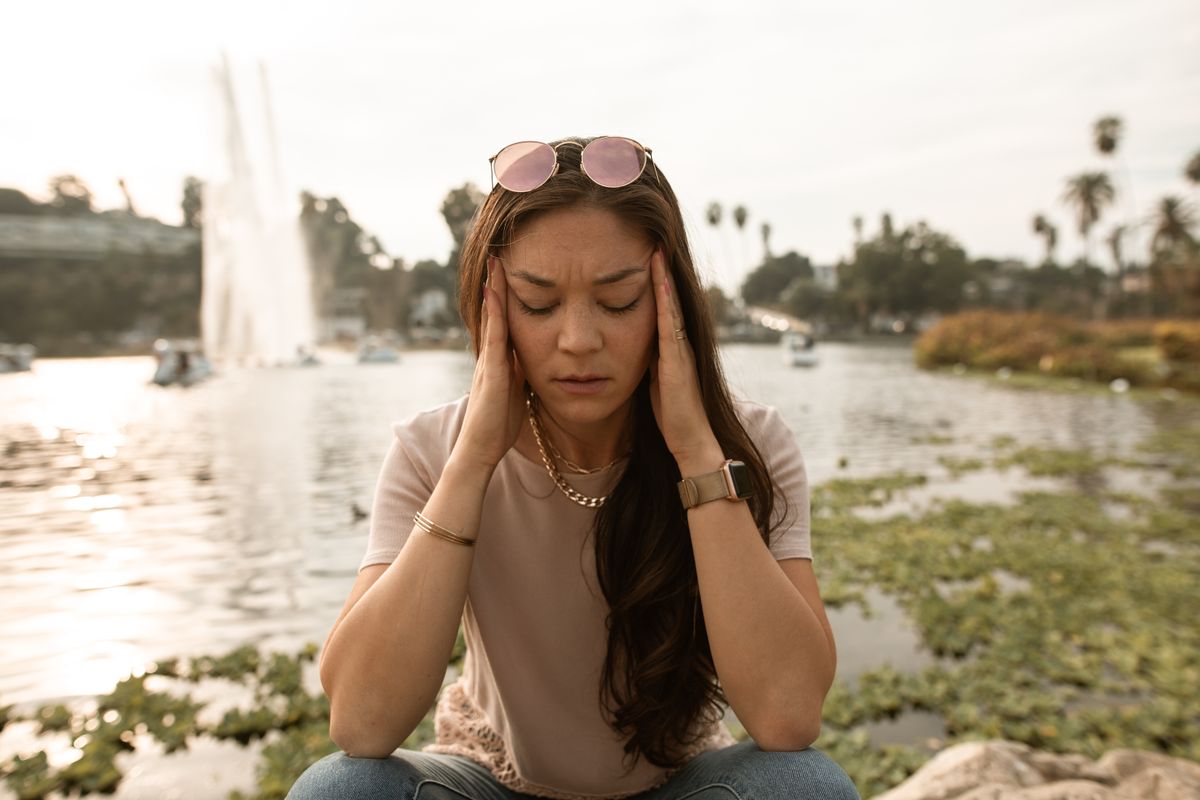 A lady sitting by a lake, visibly stressed and holding their temples.