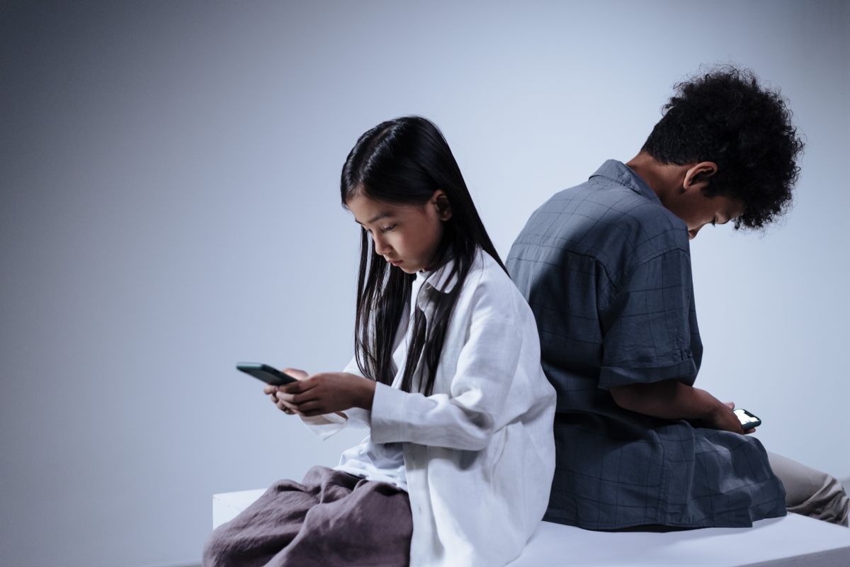 Two Teens individuals sitting back-to-back while looking down at their respective smartphones.