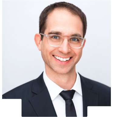 Smiling man wearing glasses, a black suit, white shirt, and black tie on light background.