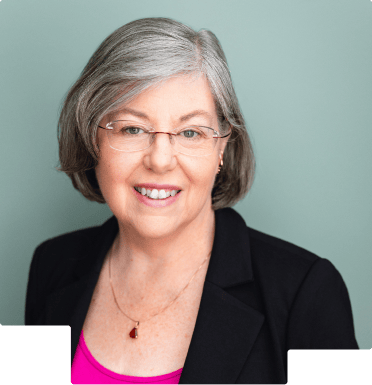 Smiling middle-aged woman with gray hair, glasses, black blazer, and pink top against a light teal background.