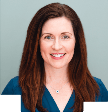 Smiling woman with long brown hair wearing a blue top and a necklace on a gray background.