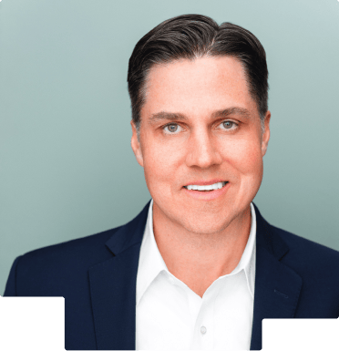 Smiling man with dark hair wearing a white shirt and dark blazer against a gray background.