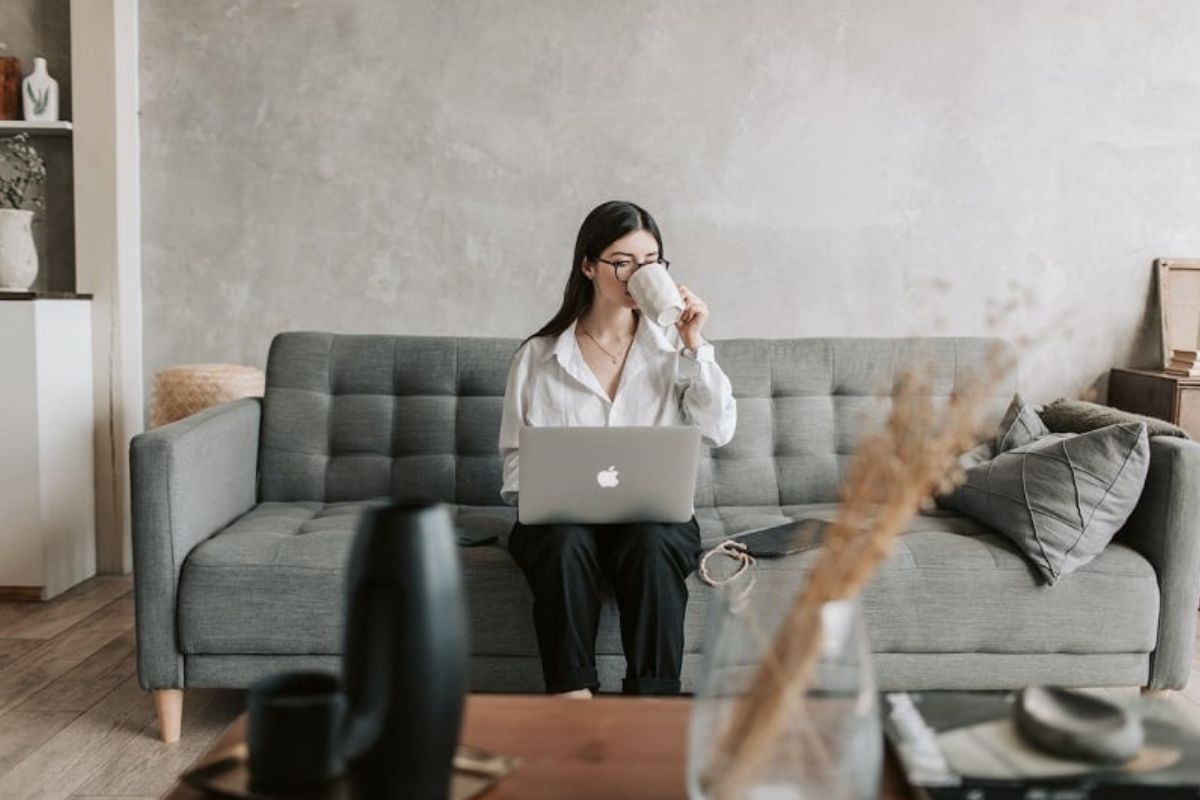 This picture shows a woman sitting on a grey couch while using a laptop.