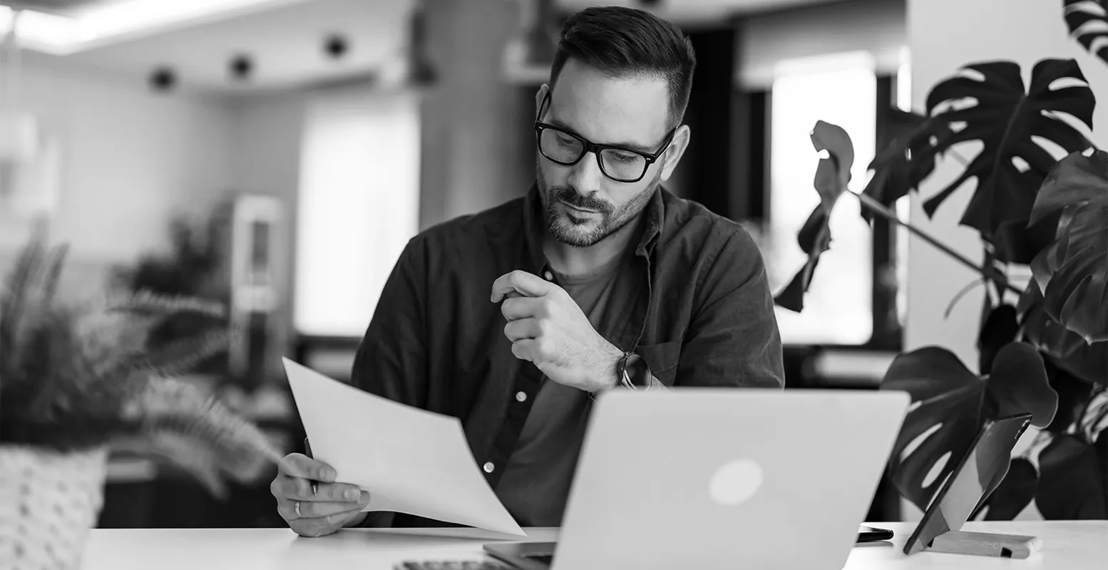 Man sitting at a desk in front of laptop looking at a sheet of paper