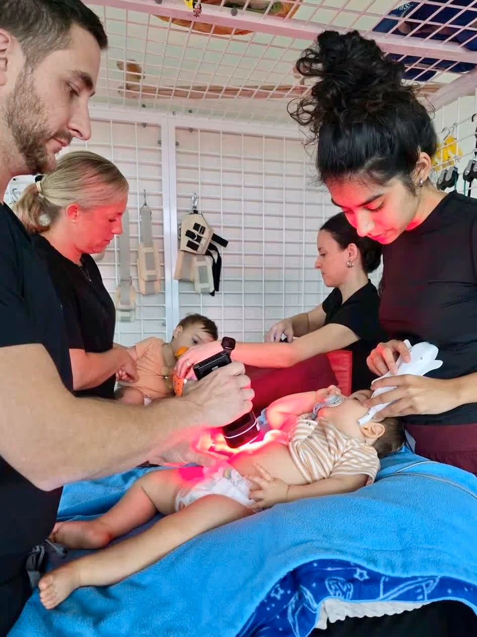 Two adults using red light therapy devices on infants lying on a blue towel during a treatment session.