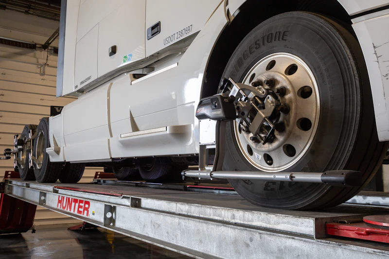 A white semi-truck is positioned on a Hunter alignment rack inside a garage, with precision alignment sensors attached to its front and rear wheels. The truck's Bridgestone tires and clean exterior contrast with minor scuff marks on the fender.