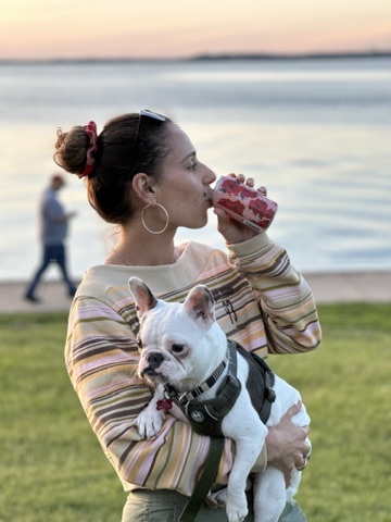 Woman with a French Bulldog holding a can, enjoying the outdoors by the water at sunset.