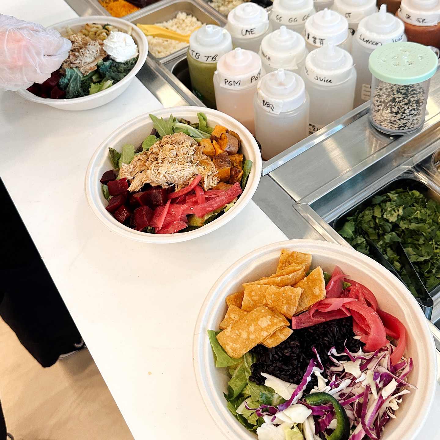 Three colorful Forage Kitchen bowls are prepared on a counter with various toppings and sauces.