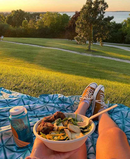 A person holds a bowl of food from Forage Kitchen on a picnic blanket with a water bottle in James Madison Park.