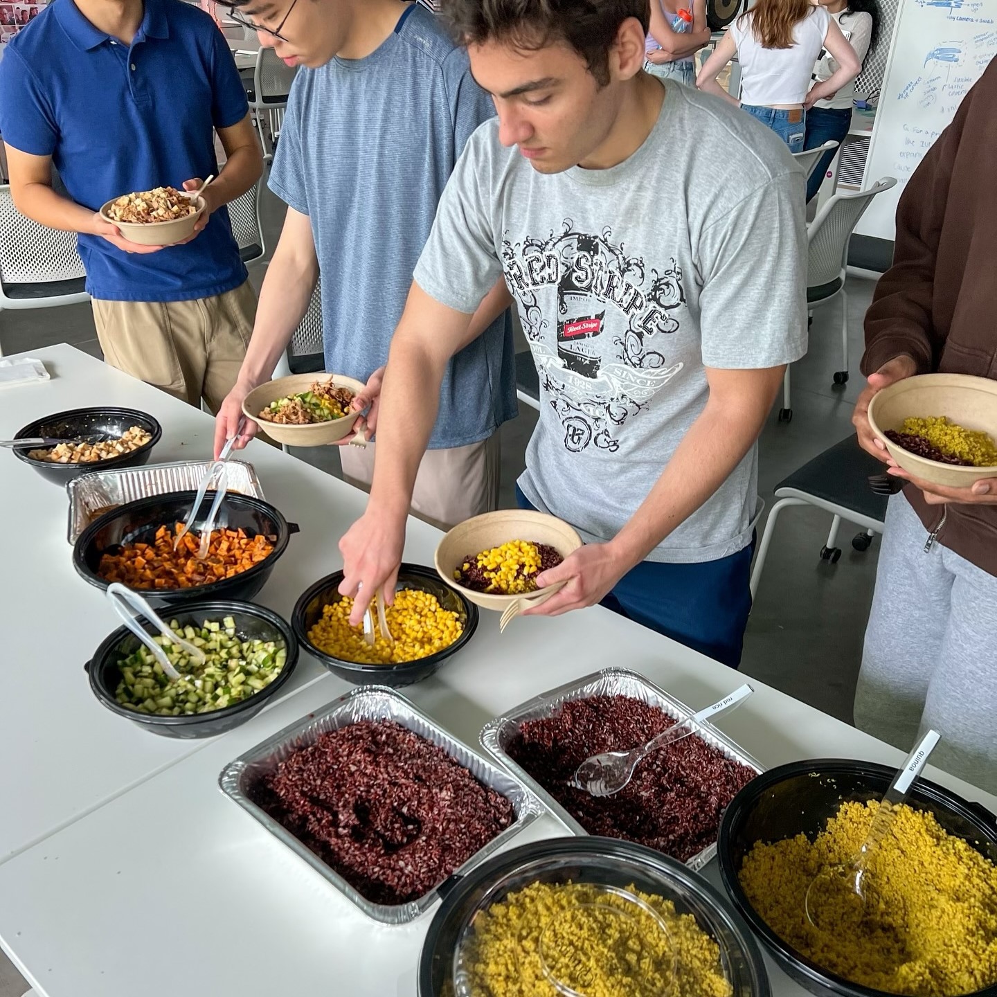A group of people filling bowls with various colorful grains and vegetables at a communal table.