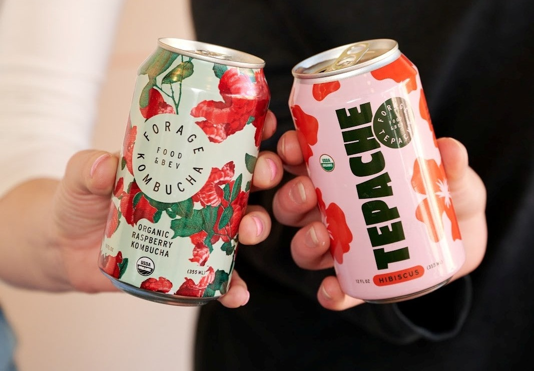 Two people, one holding a Forage Kitchen tepache can and another holding a Forage Kitchen kombucha can.