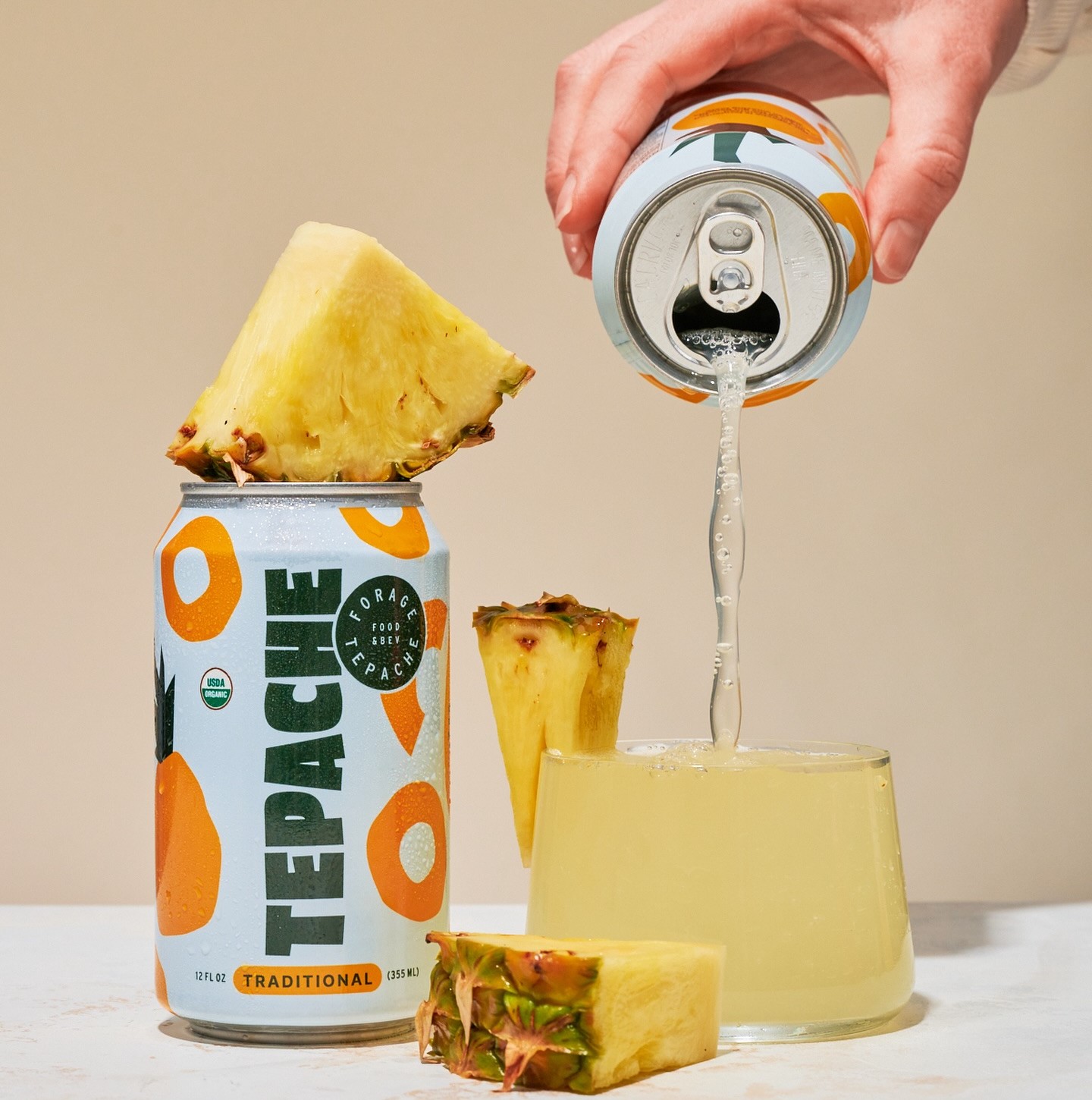  A hand pours Forage Tepache from a can into a glass, with pineapple slices around the can and glass.