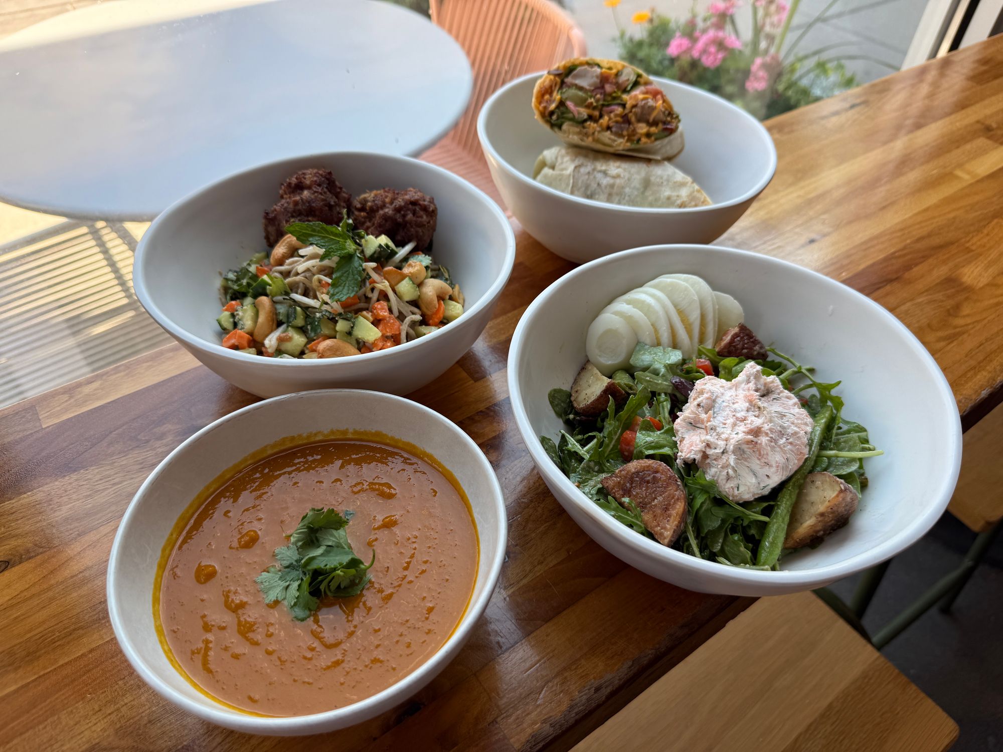 Four bowls on a wooden table featuring a grain bowl, soup, salad, and a fresh wrap from Forage Kitchen.