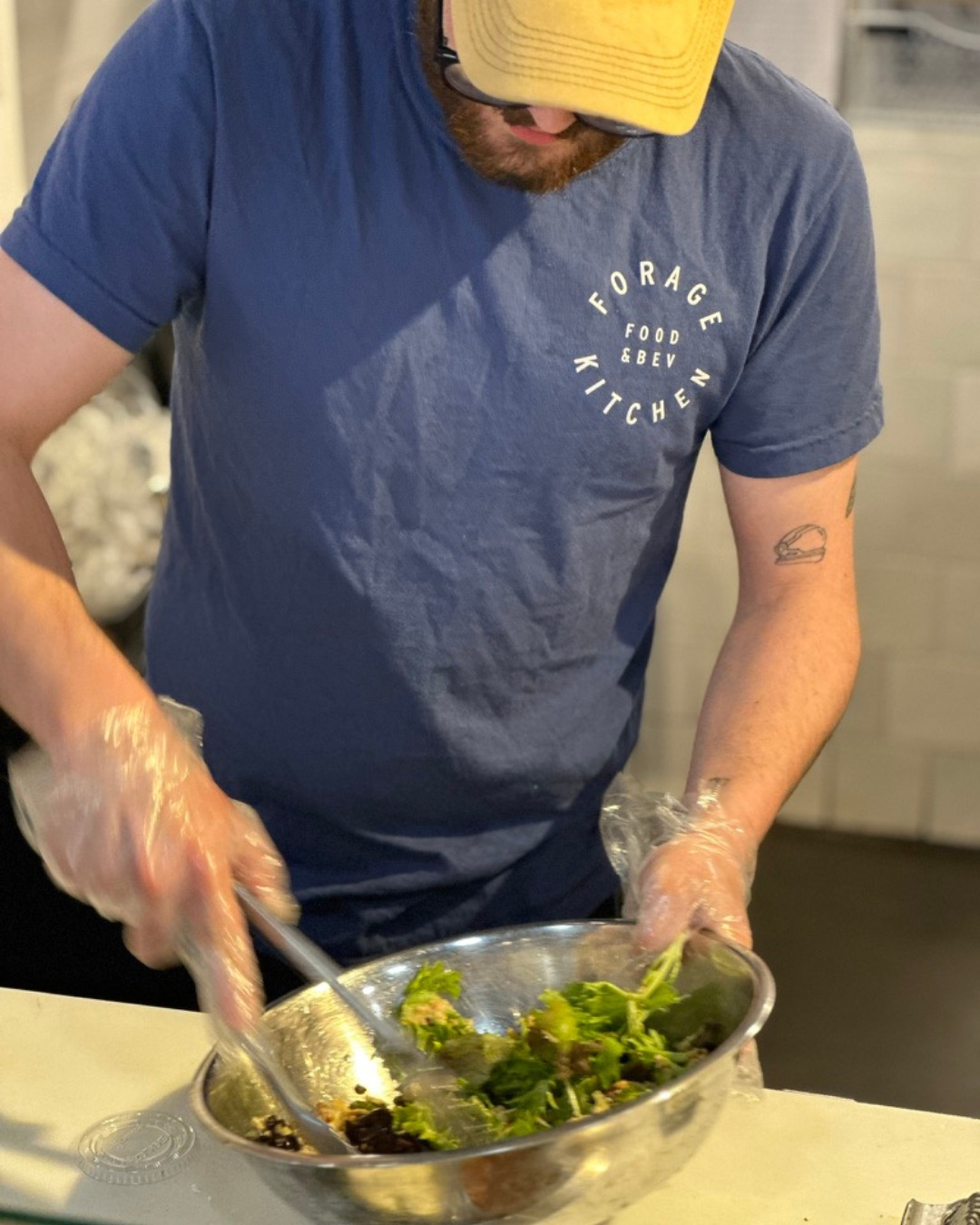 A person in a blue t-shirt and a yellow cap preparing an order in the kitchen of Forage Kitchen. 