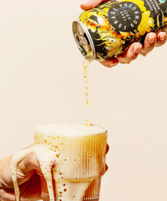 A Forage Kitchen Kombucha can pour a fizzy drink into a glass, overflowing with bubbly foam.