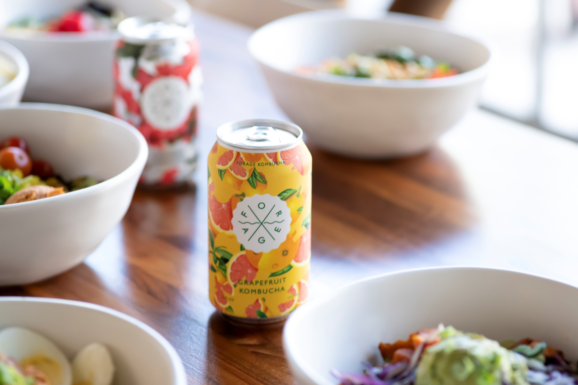 A Forage Kitchen Kombucha can on a table with salad bowls and other drinks in the background.
