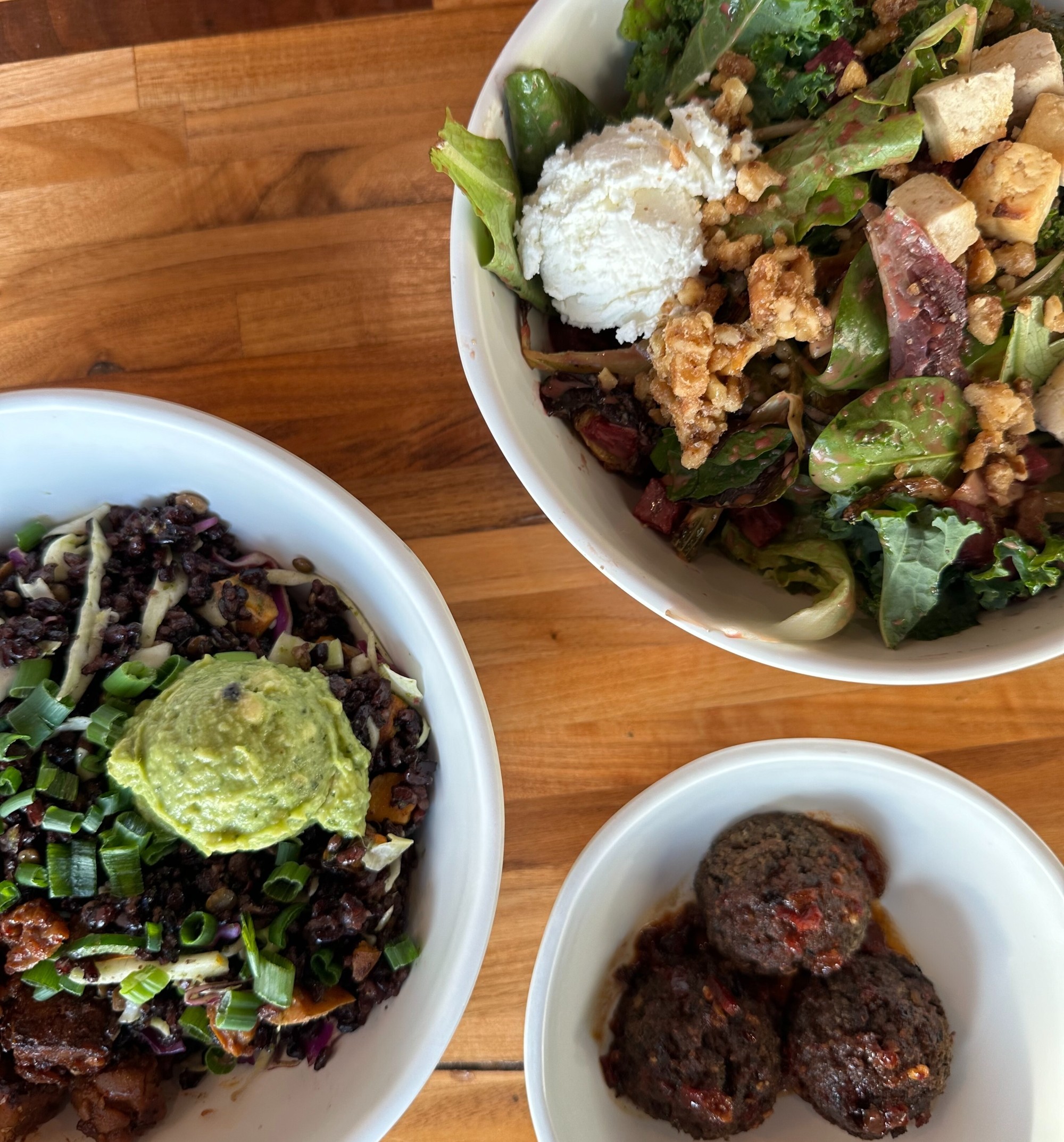 Two Forage Kitchen Salad Bowls with greens and a bowl with lentil meatballs on a table. 
