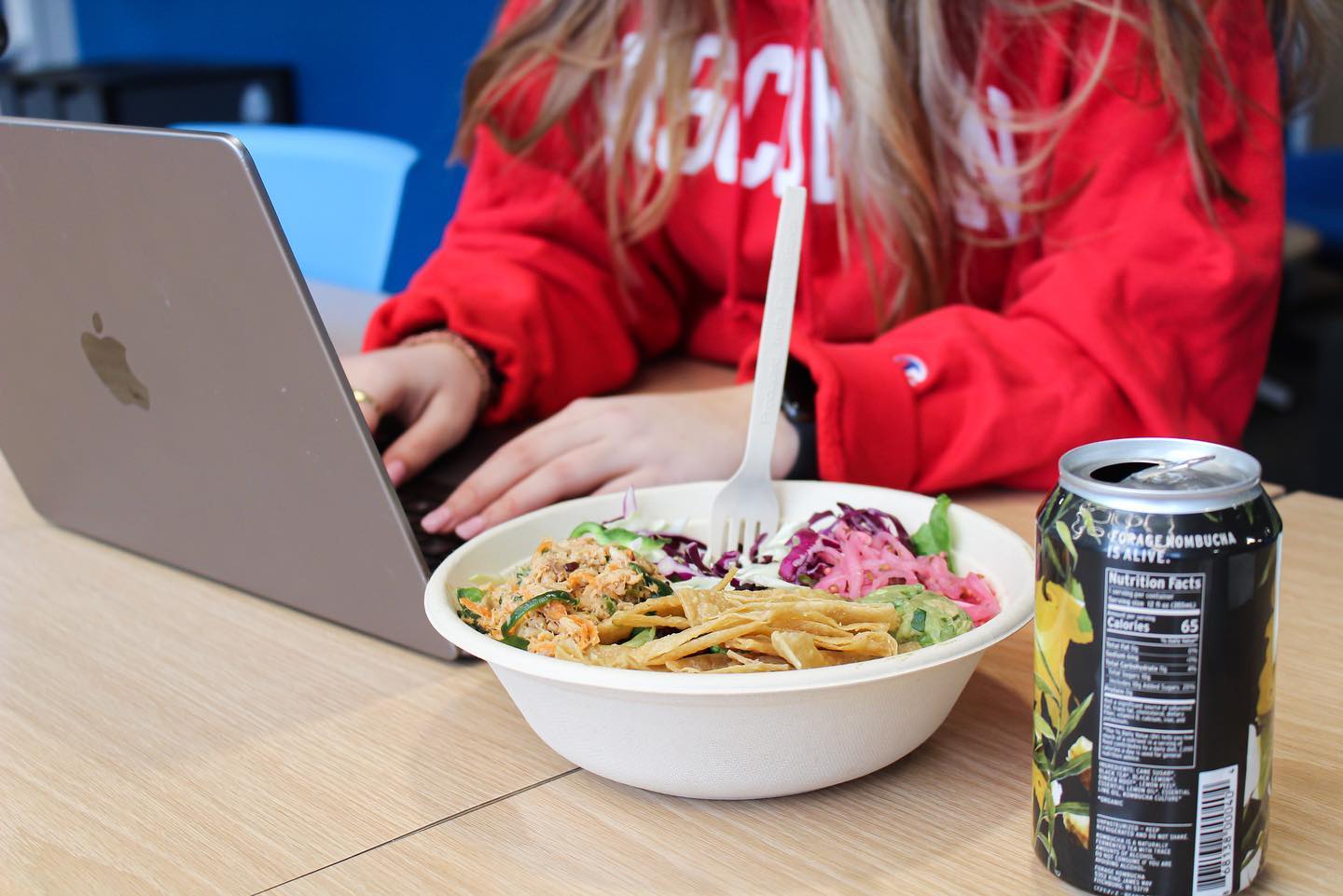 Woman in a red sweatshirt working on a laptop, enjoying Forage Kitchen’s fresh grain bowl with kombucha on the side.