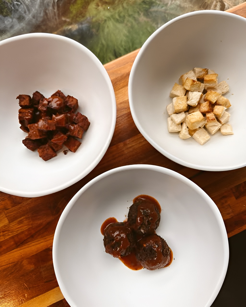 Three bowls of Forage Kitchen toppings: roasted tofu, tempeh, and saucy lentil meatballs on a wooden table.