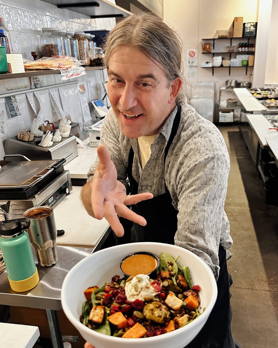Forage Kitchen chef smiles while handing a warm grain bowl to the camera inside a busy, welcoming kitchen.