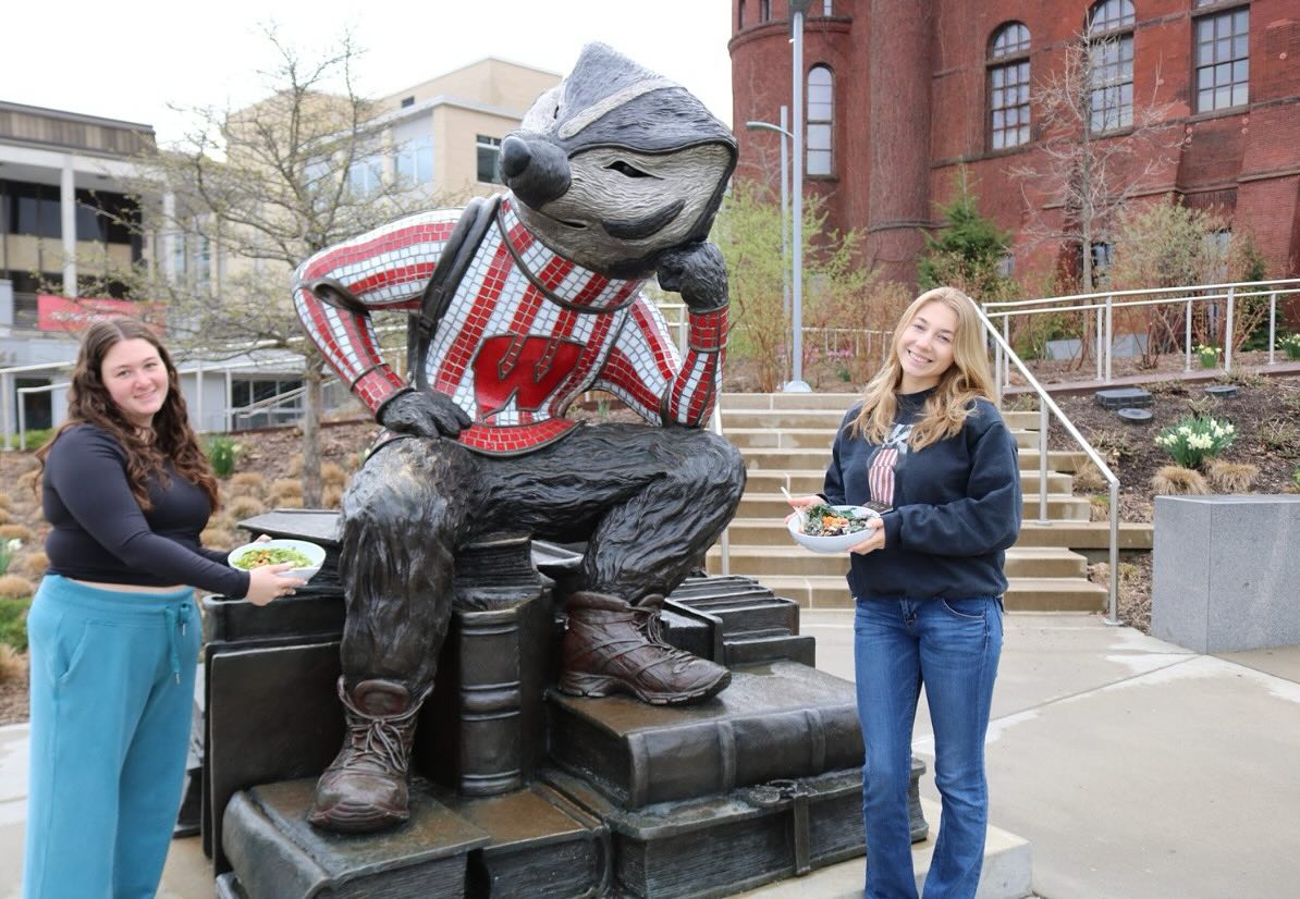 Two UW–Madison students holding Forage Kitchen bowls pose next to a statue of Bucky Badger on campus.