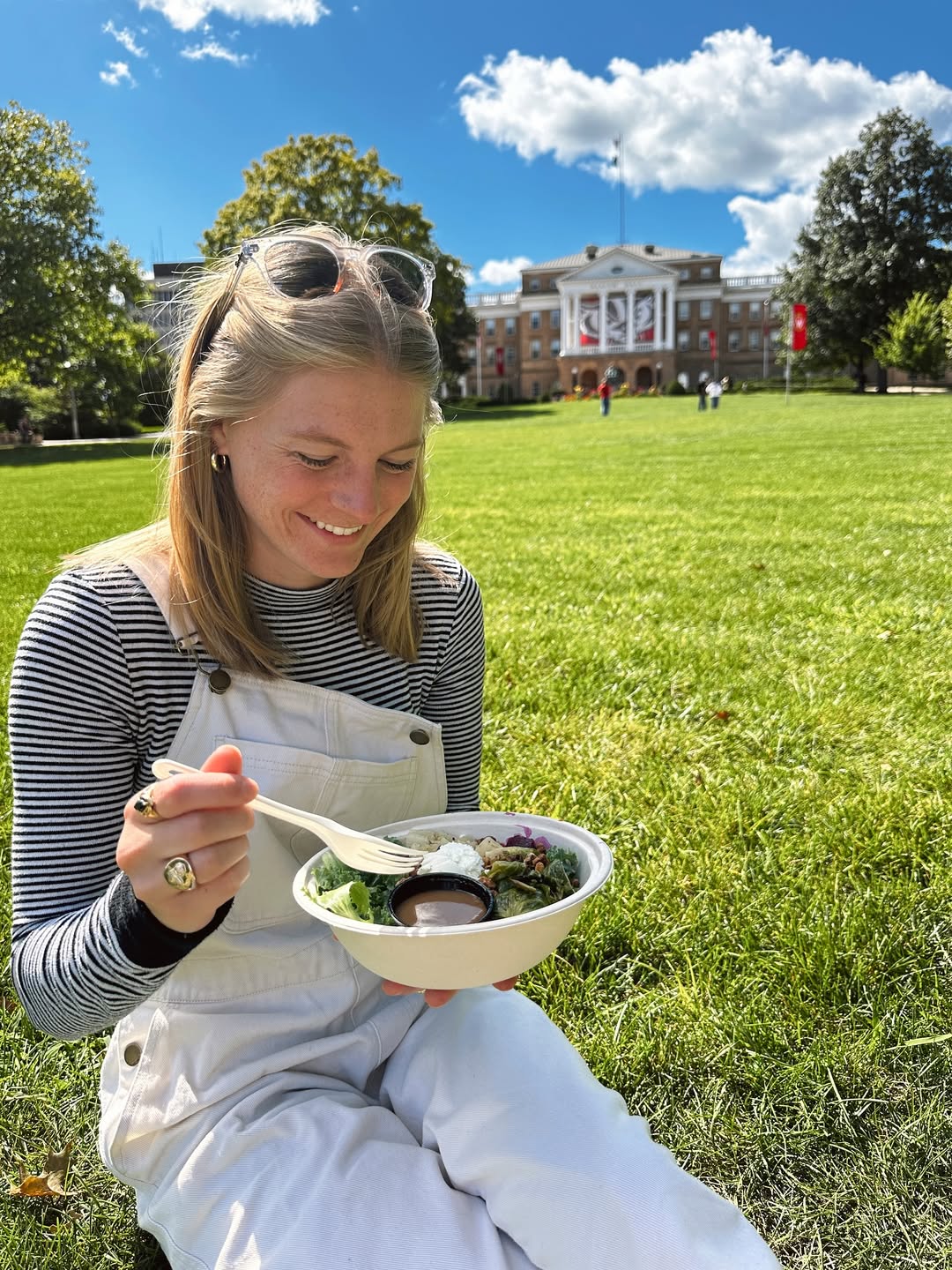 A UW–Madison student sitting on grass eating a Forage Kitchen grain bowl between classes on a sunny day.