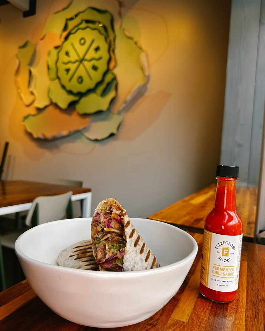 A wrap and a bottle of locally made Fermented Chili Sauce displayed on a table with the Forage Kitchen logo backdrop behind them.