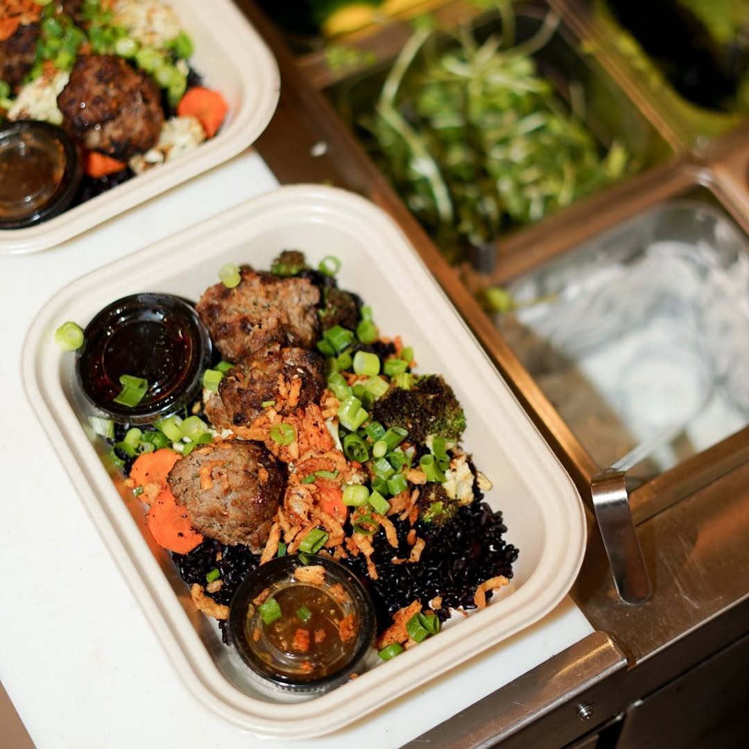 Close-up of a Forage Kitchen Japanese BBQ plate with beef meatballs, black rice, and togarashi onions on a wooden counter.
