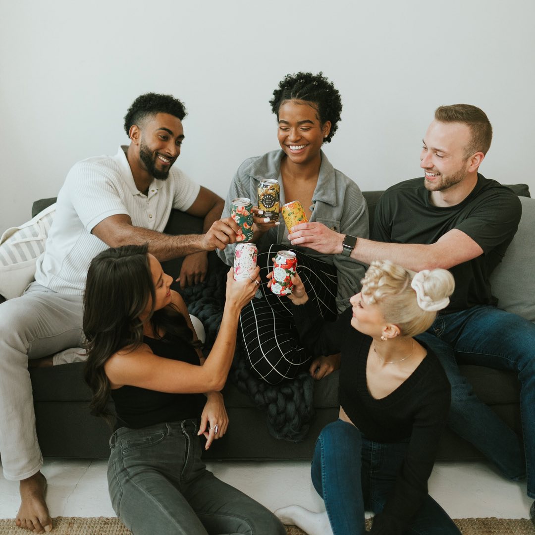 A group of friends sitting on a couch, raising cans of Forage Kombucha in a cheerful toast.