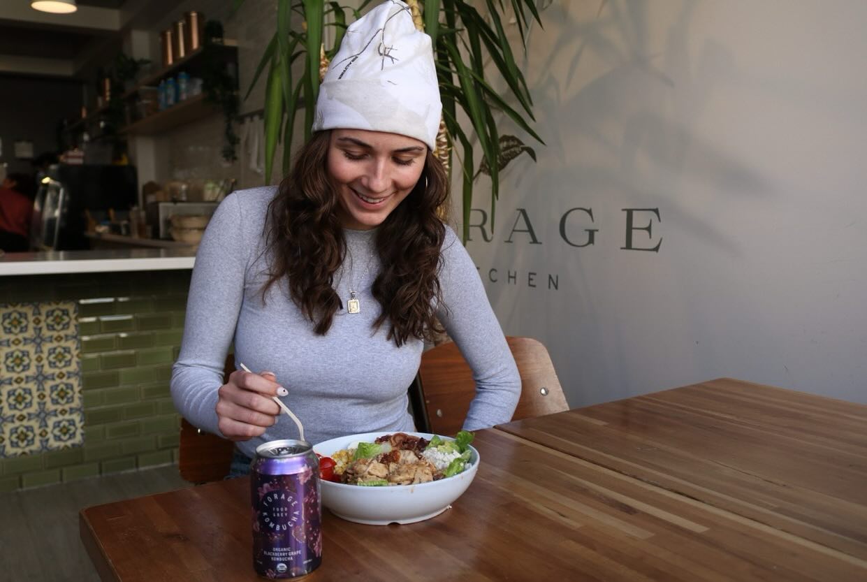 A smiling woman wearing a winter hat eating a salad at Forage Kitchen, with a kombucha bottle on the wooden table.