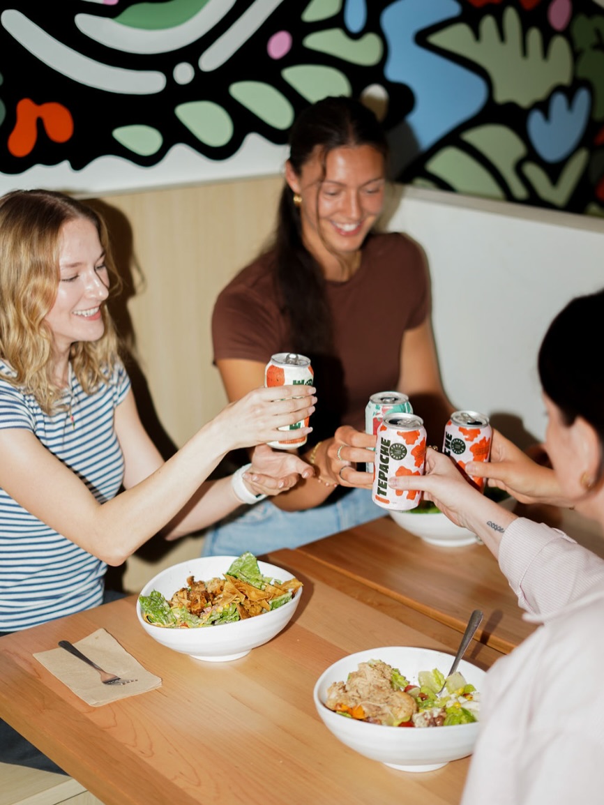  Four friends clinking kombucha cans over grain bowls at a Forage Kitchen restaurant in Whitefish Bay.
